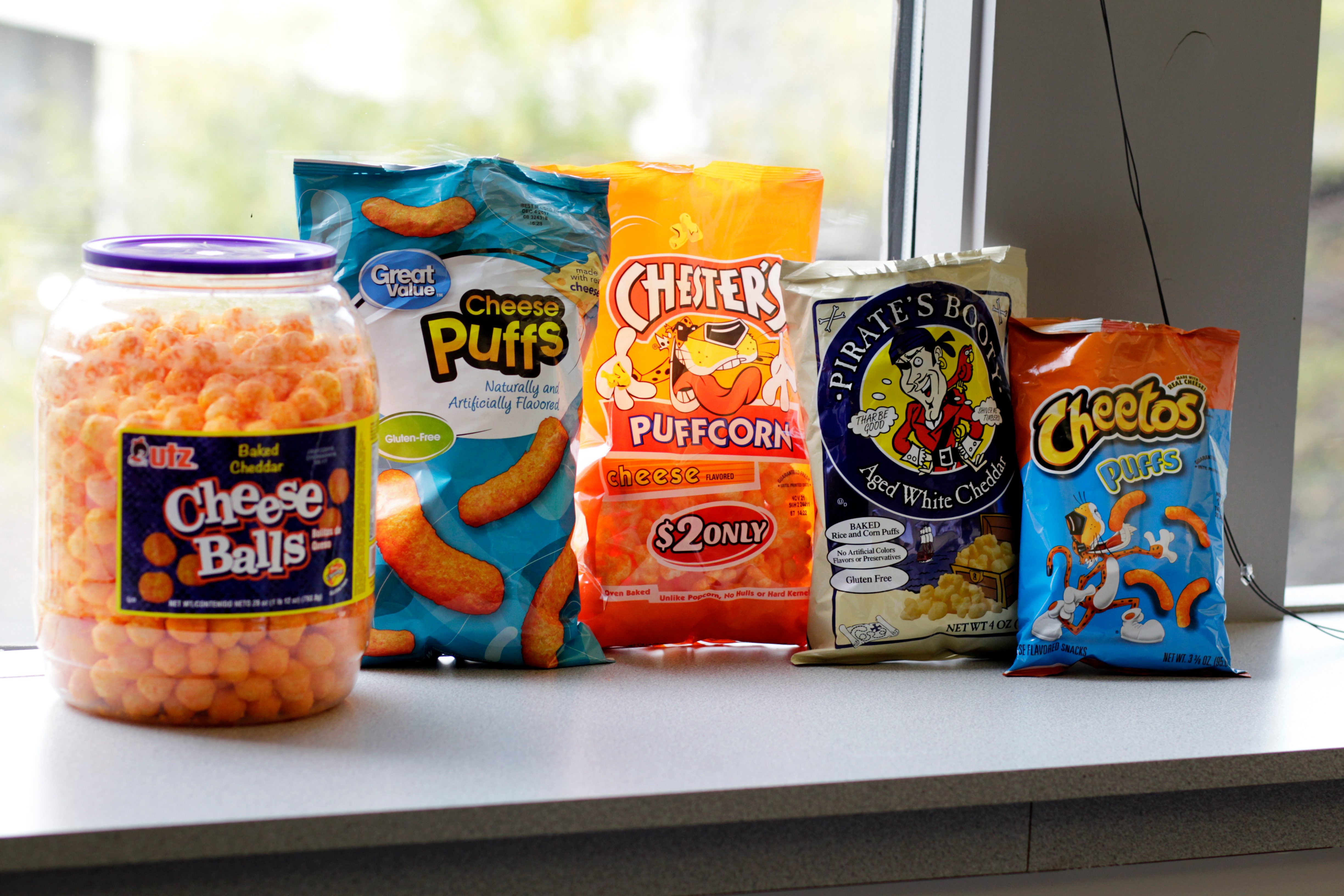 Different brands of cheese puffs lined up in front of a window