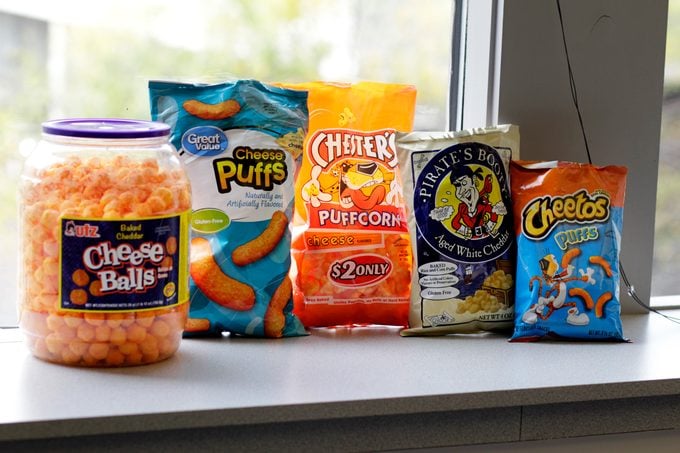 Different brands of cheese puffs lined up in front of a window