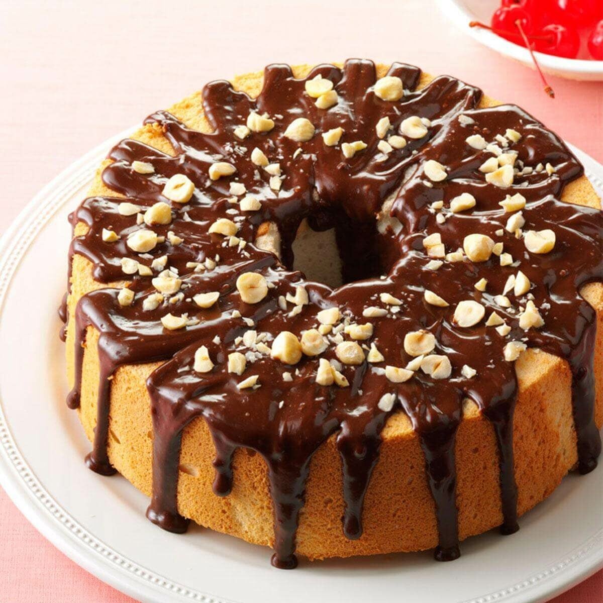 A round chiffon cake on a white plate, topped with glossy chocolate glaze and sprinkled with chopped nuts. A bowl of cherries is visible in the background.