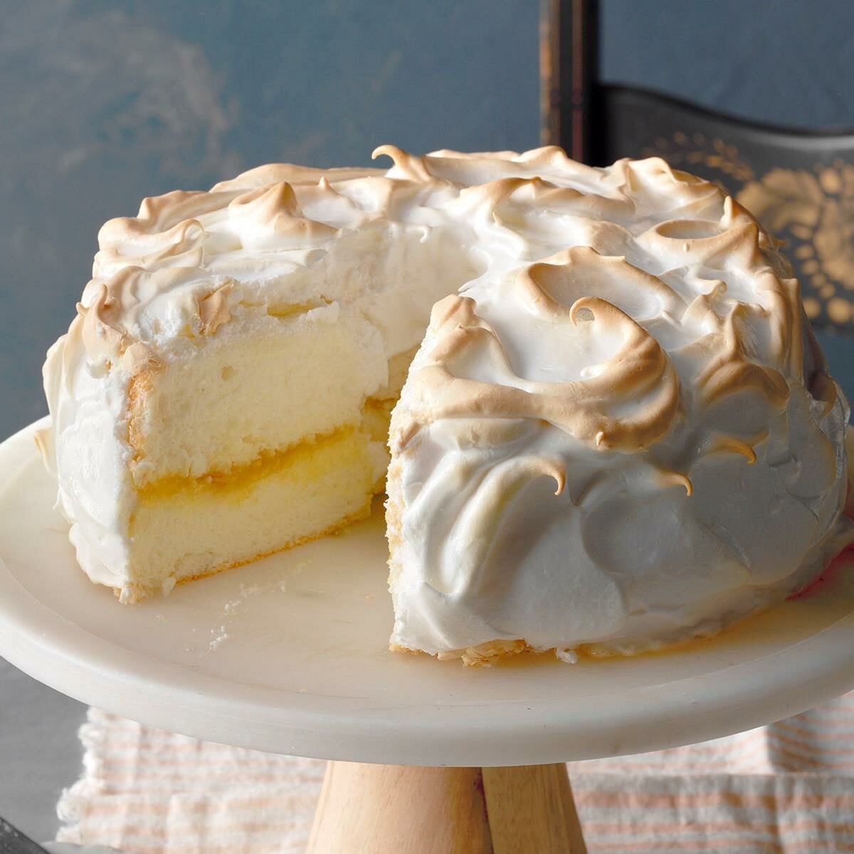A round, two-layer cake with fluffy white meringue frosting on top and a slice missing, revealing yellow filling between the layers, displayed on a white cake stand.