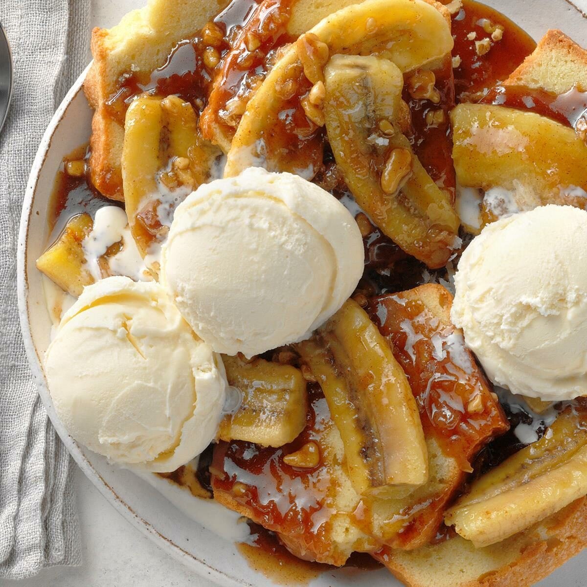 A plate of bananas foster topped with caramel sauce, chopped nuts, and three scoops of vanilla ice cream, served on a white plate next to a gray napkin.