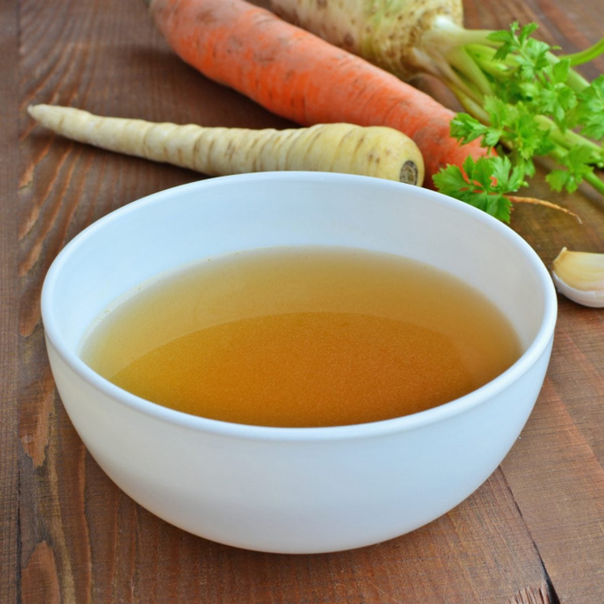 Clear beef broth, bone broth, bouillon in white bowl and vegetables on wooden table