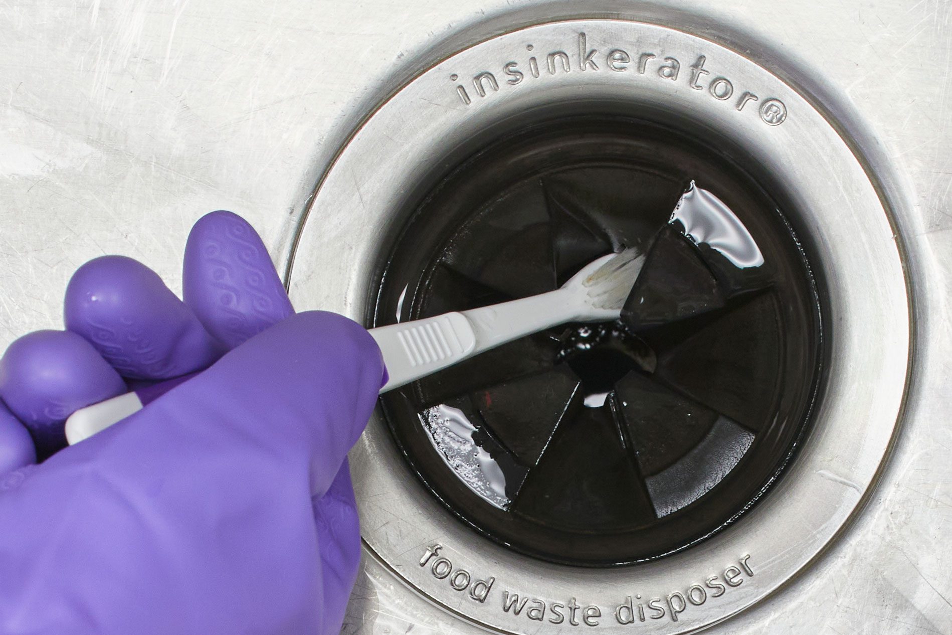 gloved hand Cleaning Garbage Disposal Splash Guard in a stainless steel sink with an old toothbrush