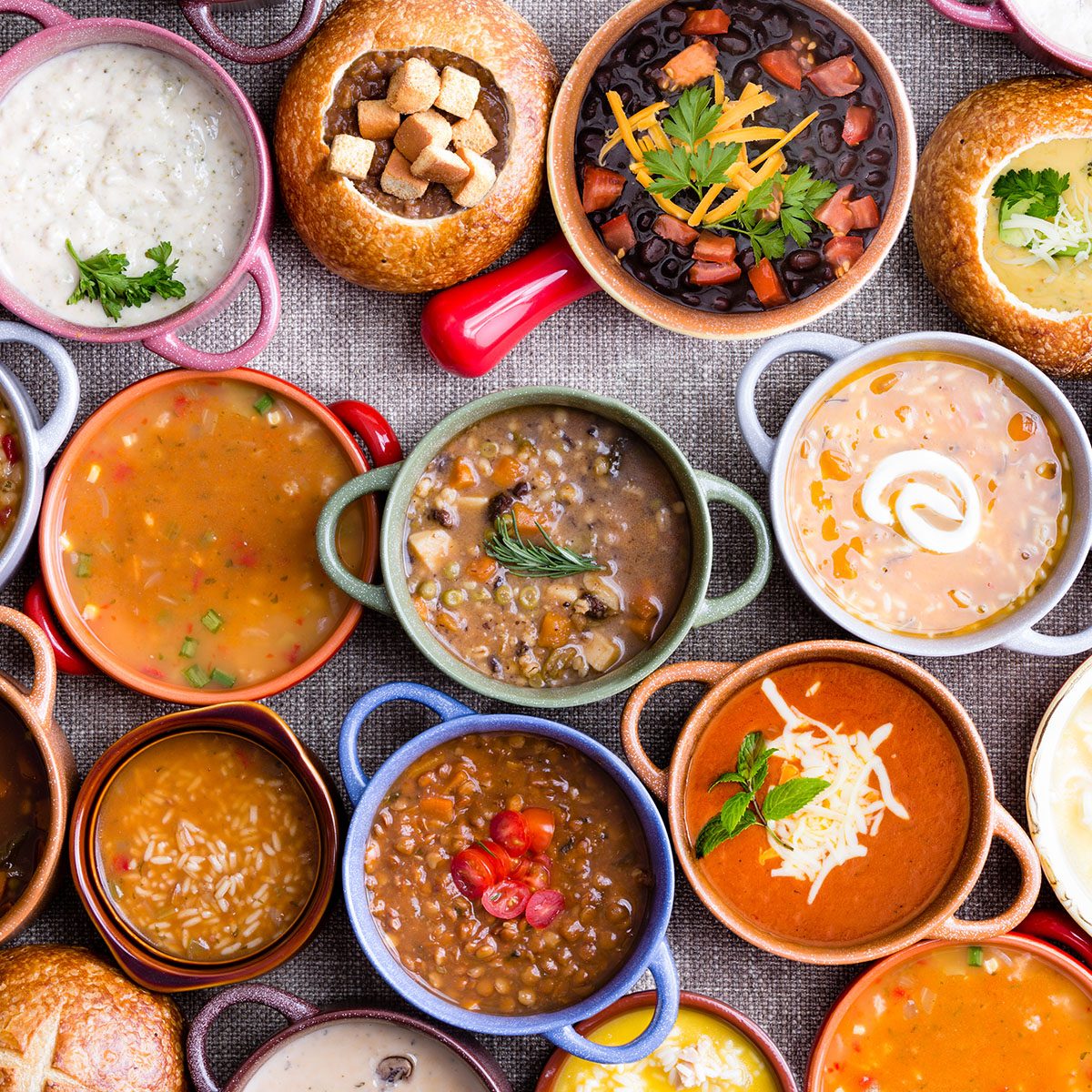 High Angle View of Various Comforting and Savory Gourmet Soups Served in Bread Bowls and Handled Dishes and Topped with Variety of Garnishes on Table Surface with Gray Tablecloth