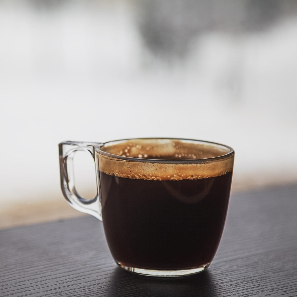 Transparent cup of coffee placed on dark brown laminated wooden surface in front of a window
