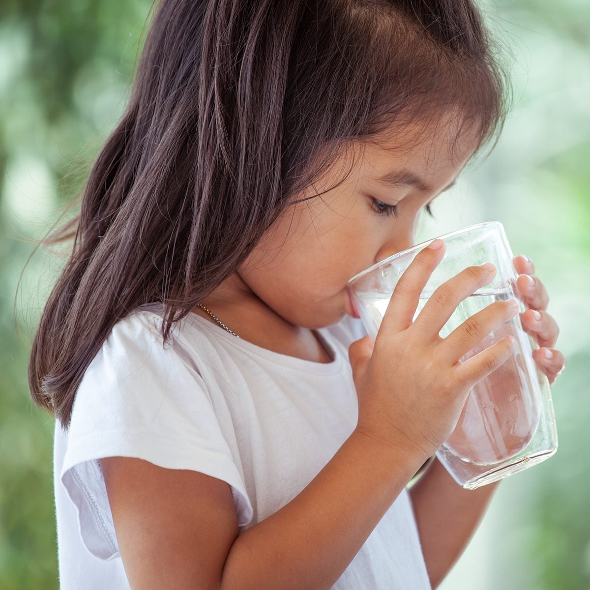 Cute asian little child girl drinking fresh water from glass on green nature background