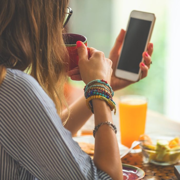 Woman using cellphone and laptop inside house and drinking coffee / tea.