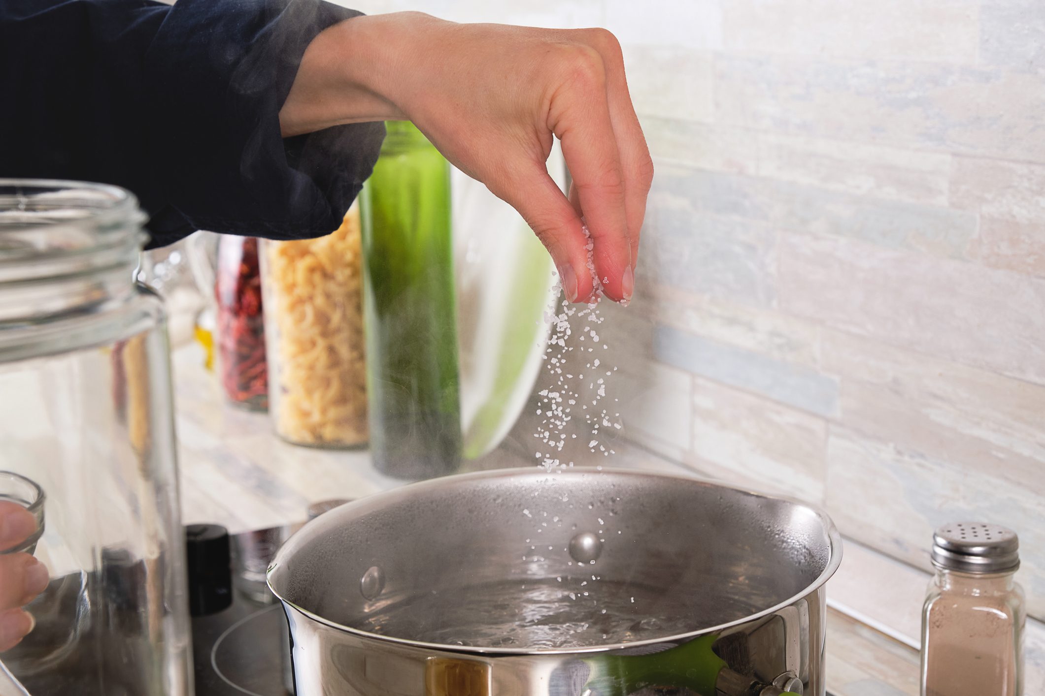 Cooking Italian pasta with cream sauce and Basil, at home. A girl or woman Adds Salt to Boiling Water. Next to it, raw Macaroni In a Glass Jar. vegetarian food. Step-by-step instructions, do it yourself. Step 2.