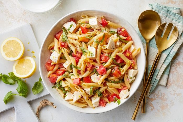 Caprese Pasta Salad in a Large Ceramic Bowl with Sliced Lemon and Basil on Left and Bronze Mixing Spoon and Pasta Server on Right