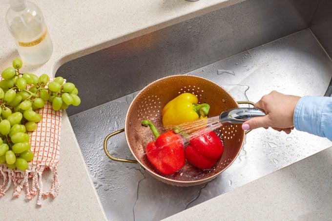three bell peppers are in a colander in a stainless steel sink. a hand uses the sprayer faucet to rinse the them..