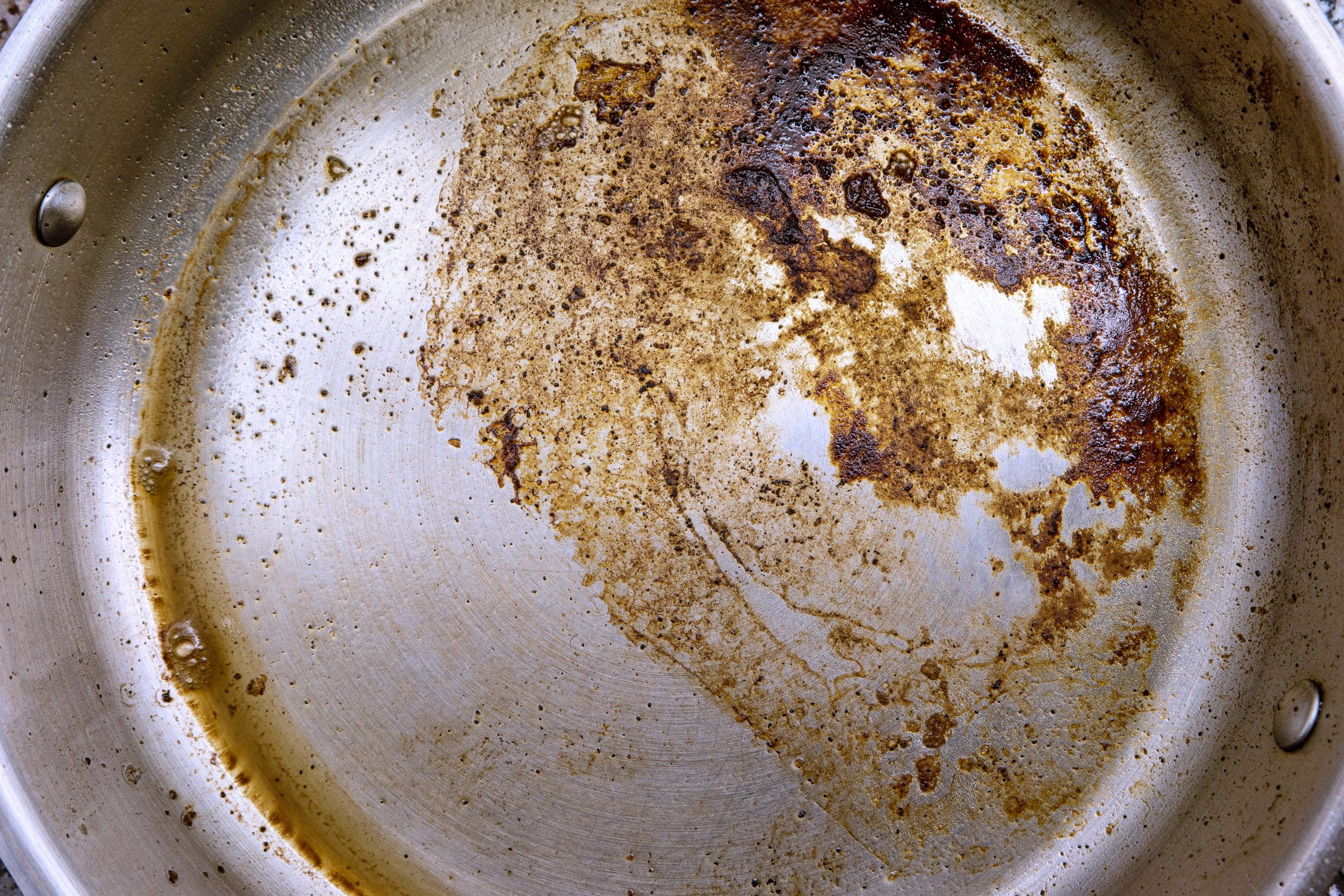 Burnt oil and fat remains on the bottom of a stainless steel frying skillet pan close up top view.
