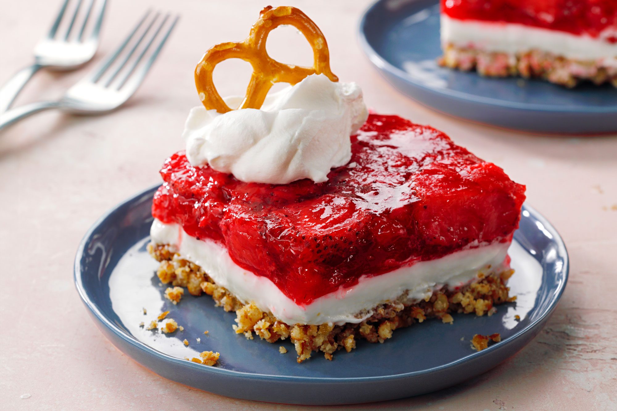 A slice of layered dessert with a pretzel crust, creamy white middle, and strawberry topping, garnished with whipped cream and a mini pretzel, served on a blue plate. Two forks are in the background.