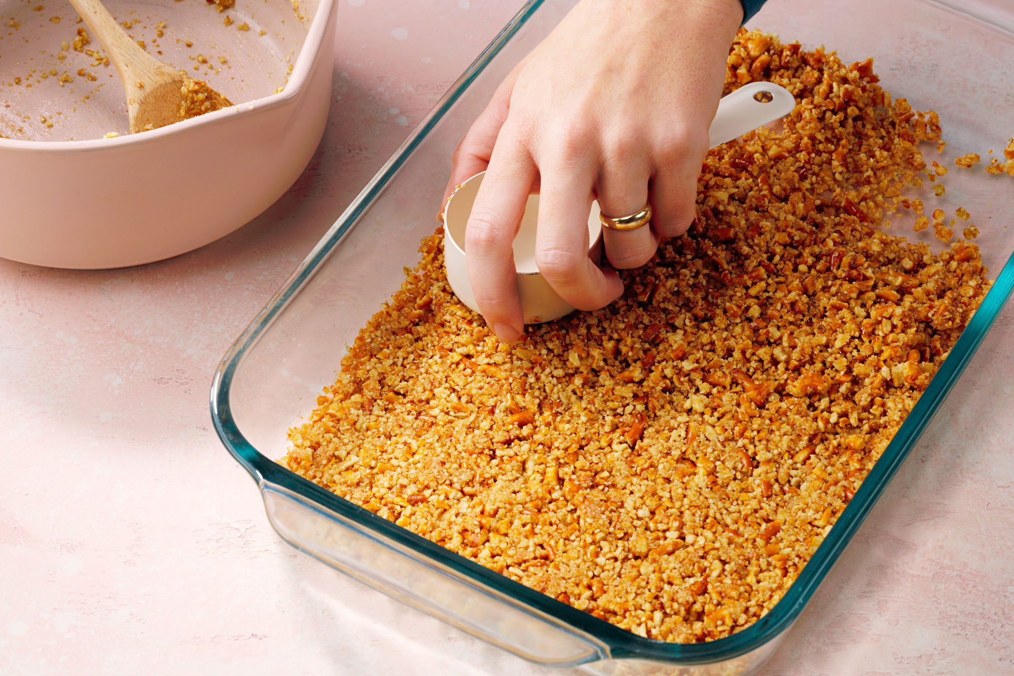 A hand presses down a layer of crushed graham crackers in a glass baking dish, using a measuring cup. A mixing bowl with a wooden spoon is beside the dish on a light pink surface.