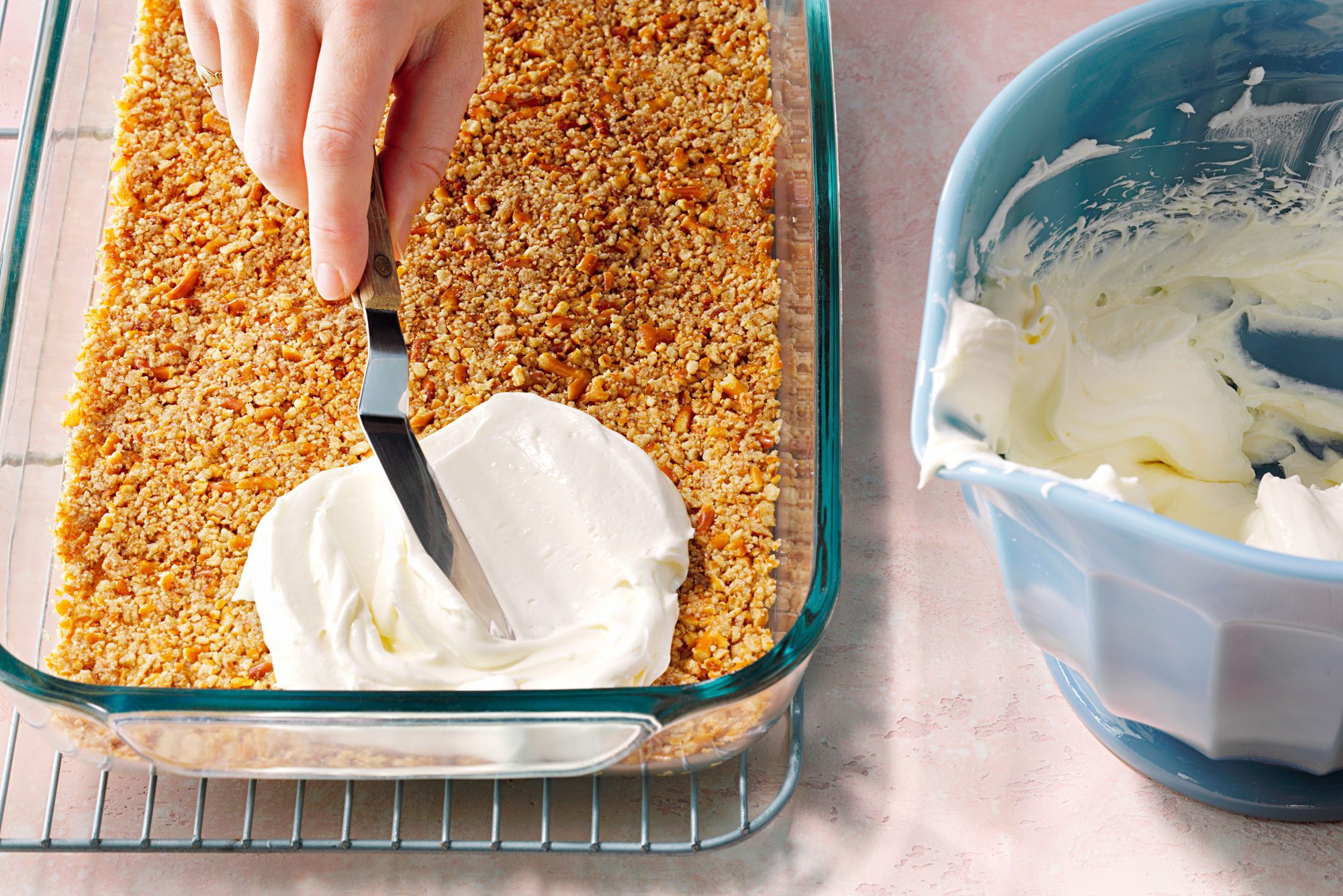 A hand spreads a layer of white frosting over a crumbly dessert in a glass baking dish, with a blue bowl of frosting beside it on a wire rack.