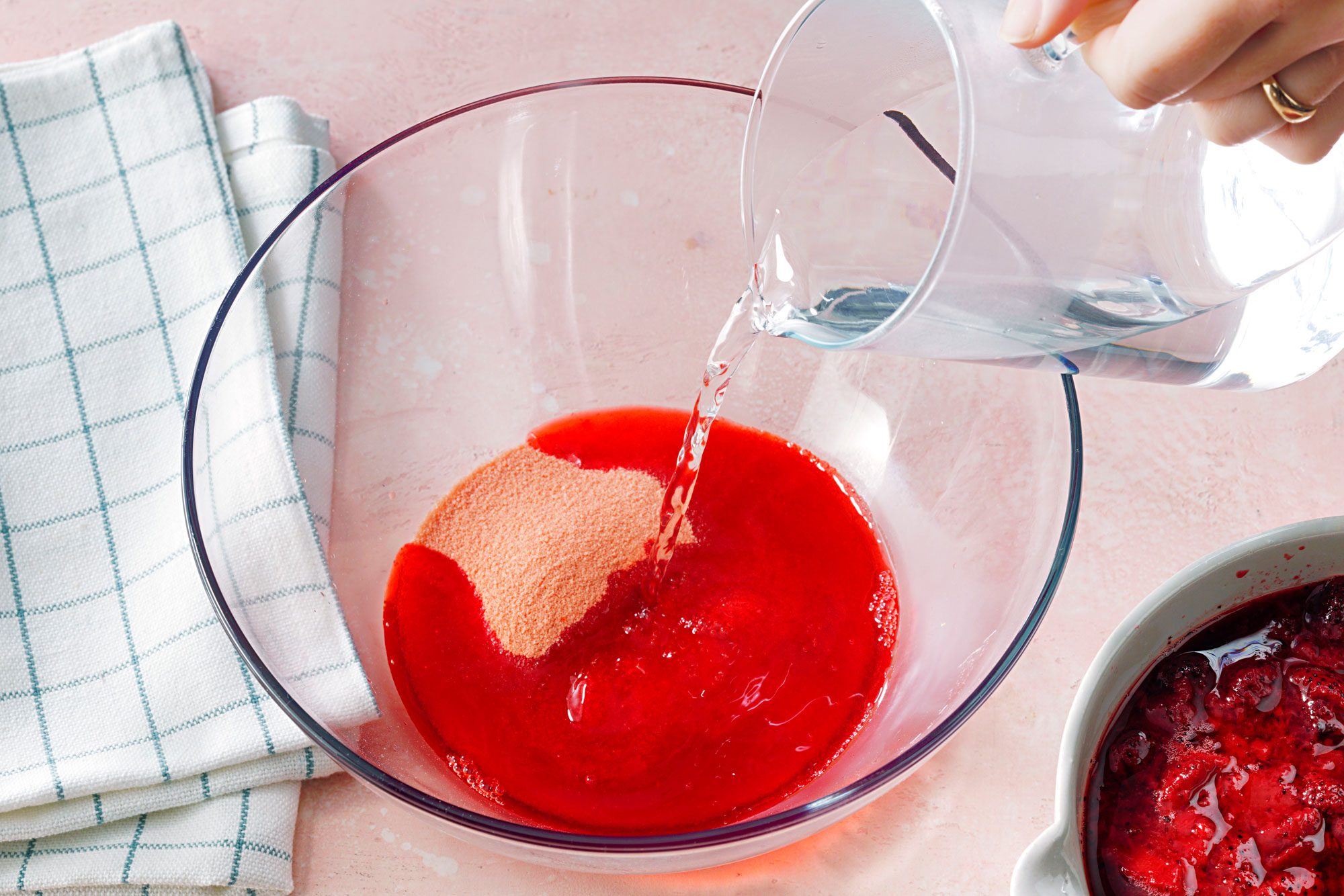 A hand pours water from a clear pitcher into a glass bowl containing red gelatin powder on a pink surface, next to a white checkered cloth and a bowl of strawberries.