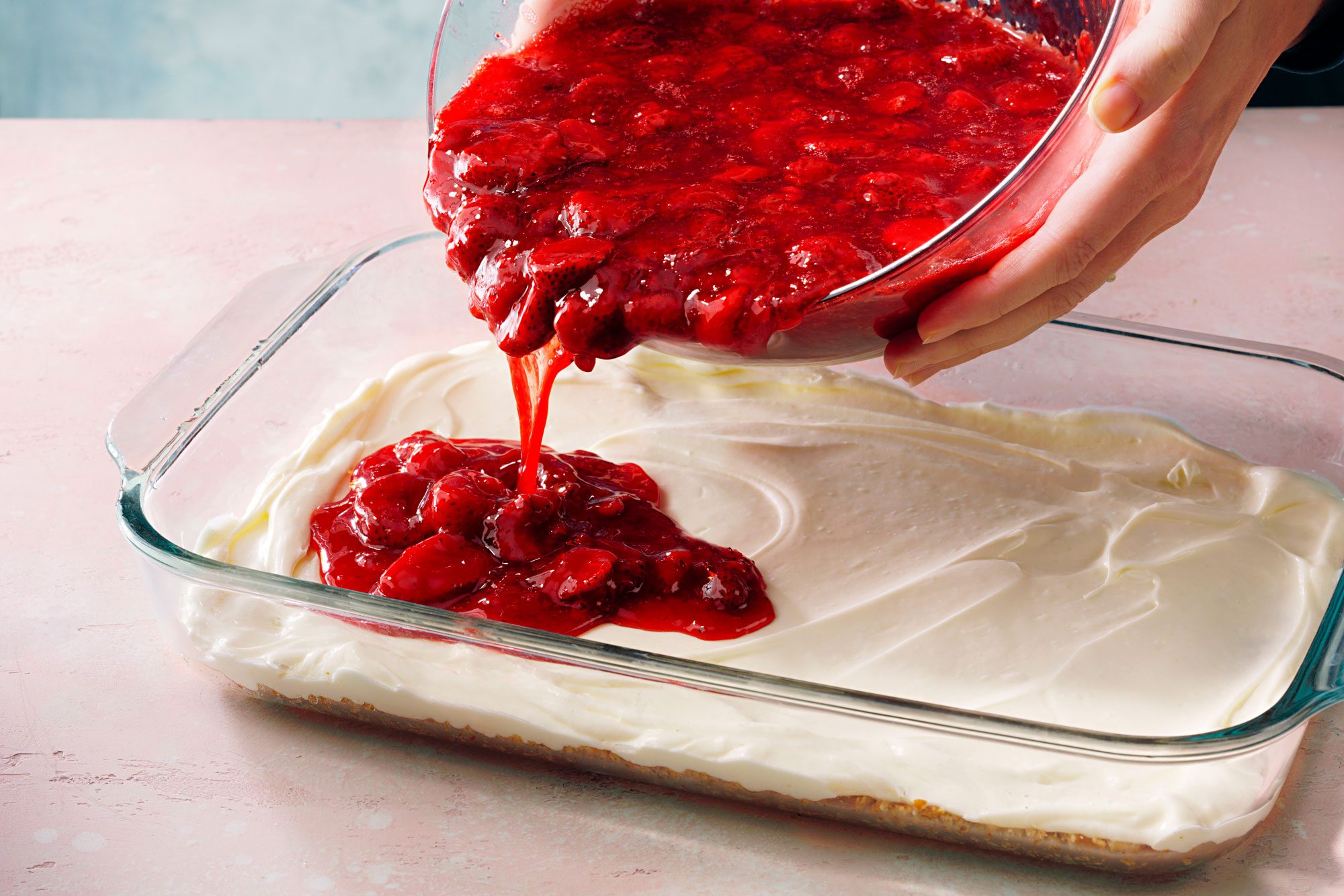 A person pours a chunky red strawberry topping over a creamy white layer in a glass baking dish, likely preparing a layered dessert on a light pink surface.