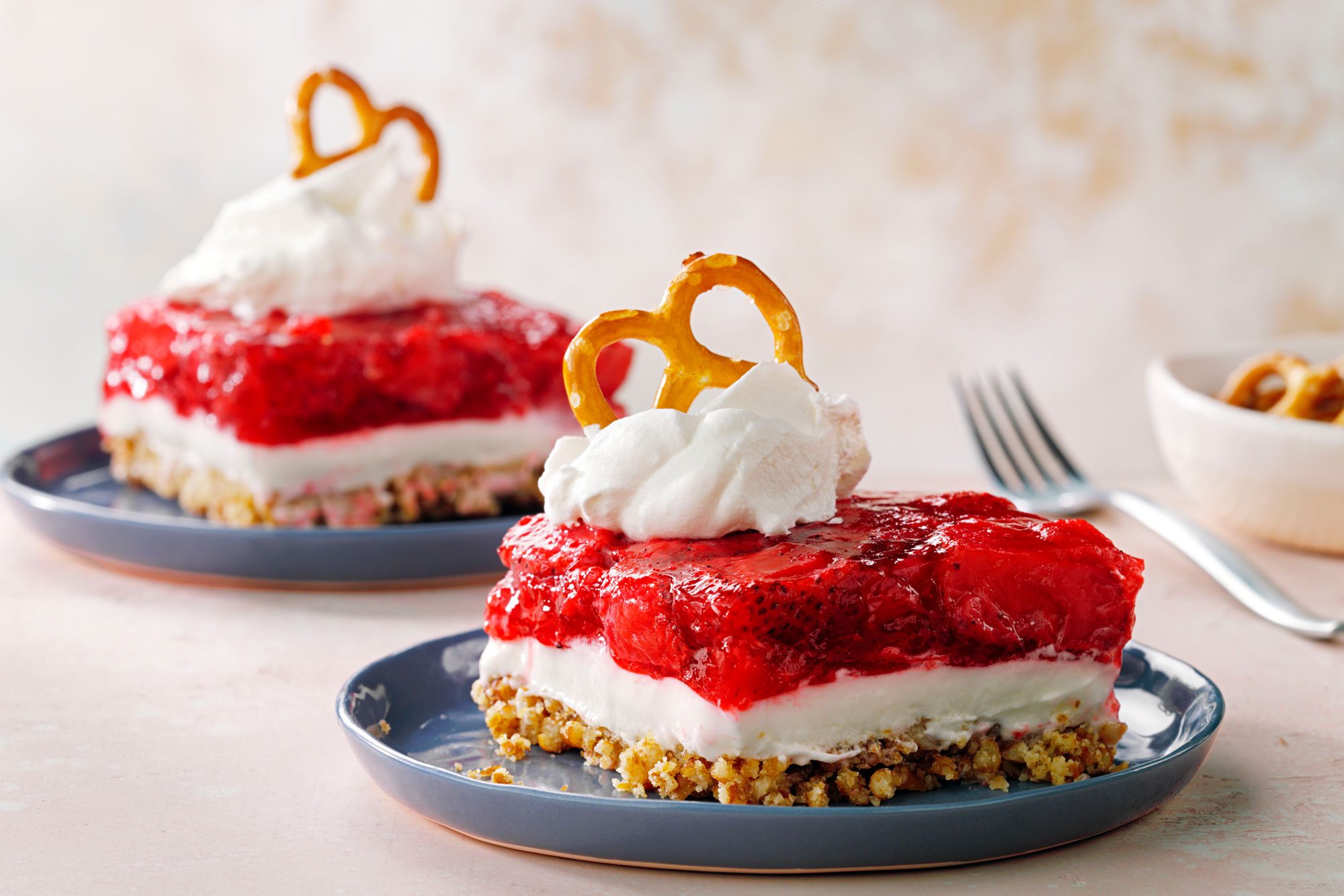 Two slices of layered strawberry pretzel dessert topped with whipped cream and a pretzel sit on blue plates. The dessert has a pretzel crust, creamy middle layer, and strawberry gelatin topping. A fork and bowl of pretzels are in the background.