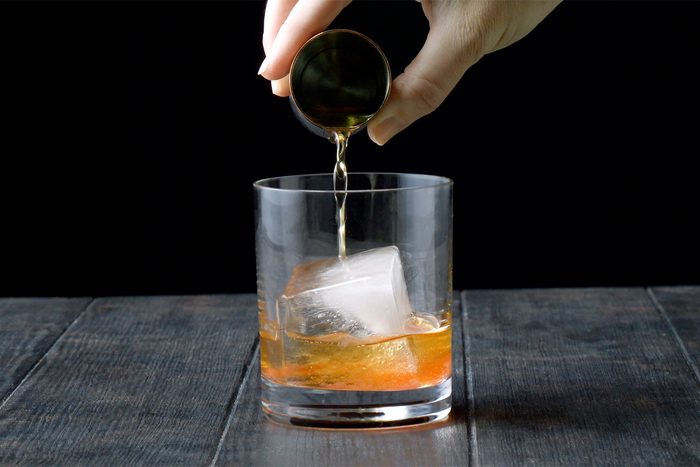 A hand is pouring a drink, possibly whiskey, from a small jigger into a glass.