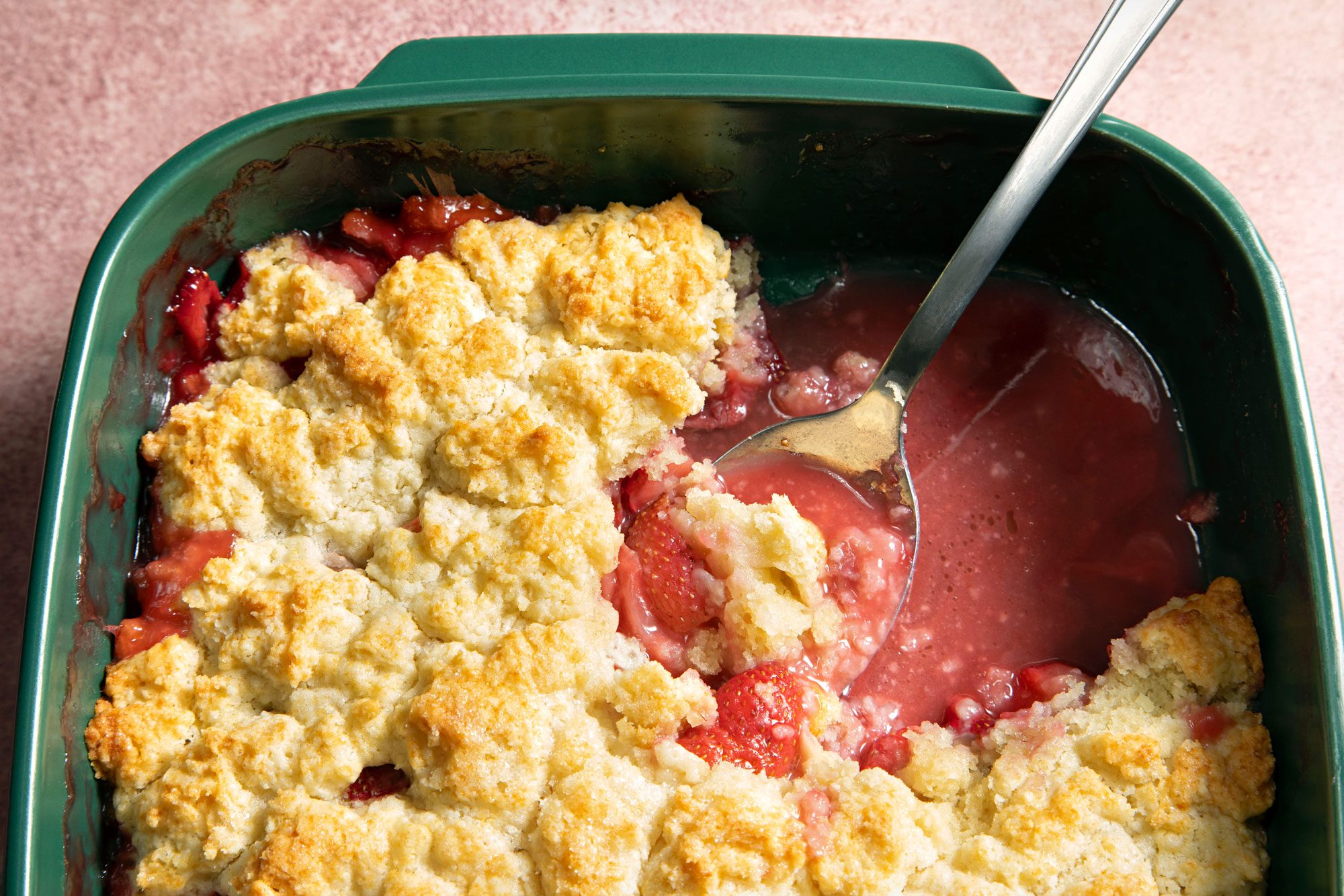 Strawberry Cobbler with Spoon in a Green Baking Dish on a Pink Surface