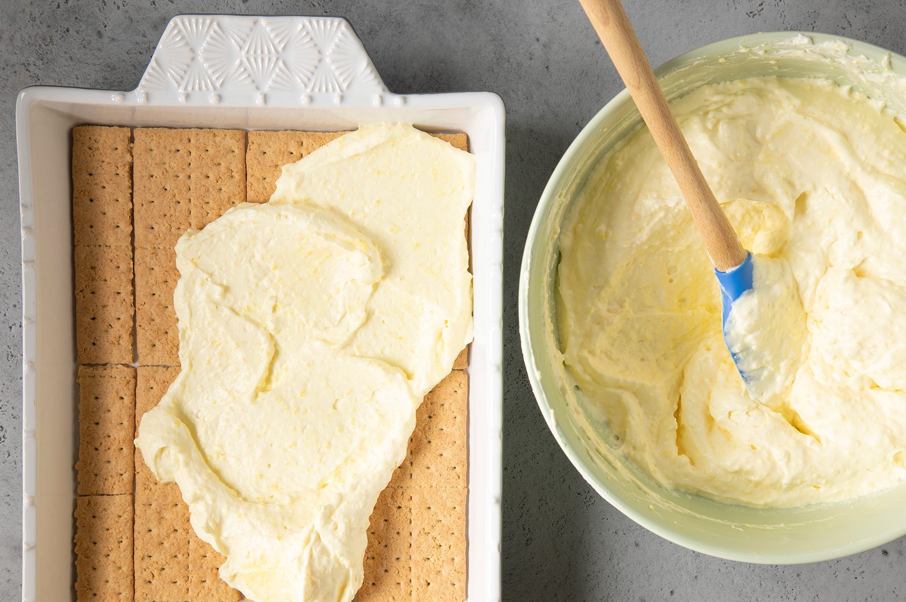 A rectangular white baking dish with a layer of graham crackers being topped with a creamy, yellow mixture.