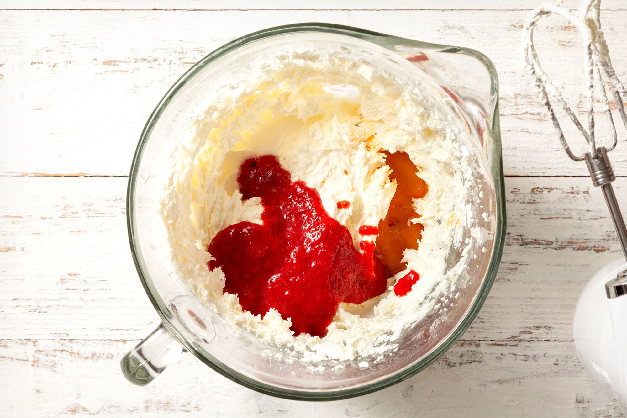 overhead shot of beaten butter and strawberry puree in a beaker with electric beater placed on right side on white wooden background