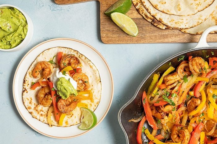 A top view of a shrimp fajita setup. A fajita with shrimp, bell peppers, sour cream, and guacamole on a plate is visible.