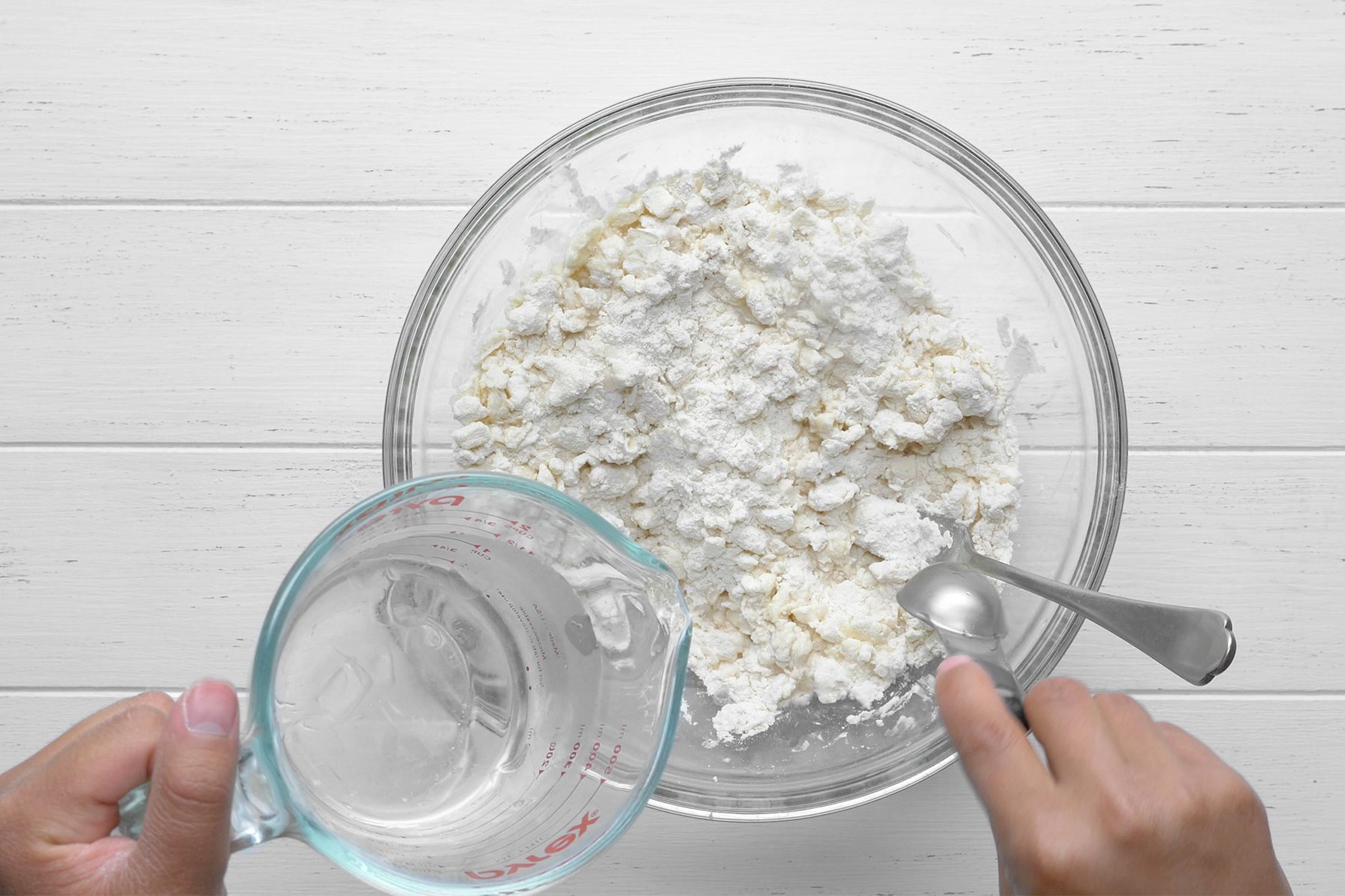 A person is adding water from a measuring jug to a glass bowl containing flour and other ingredients.