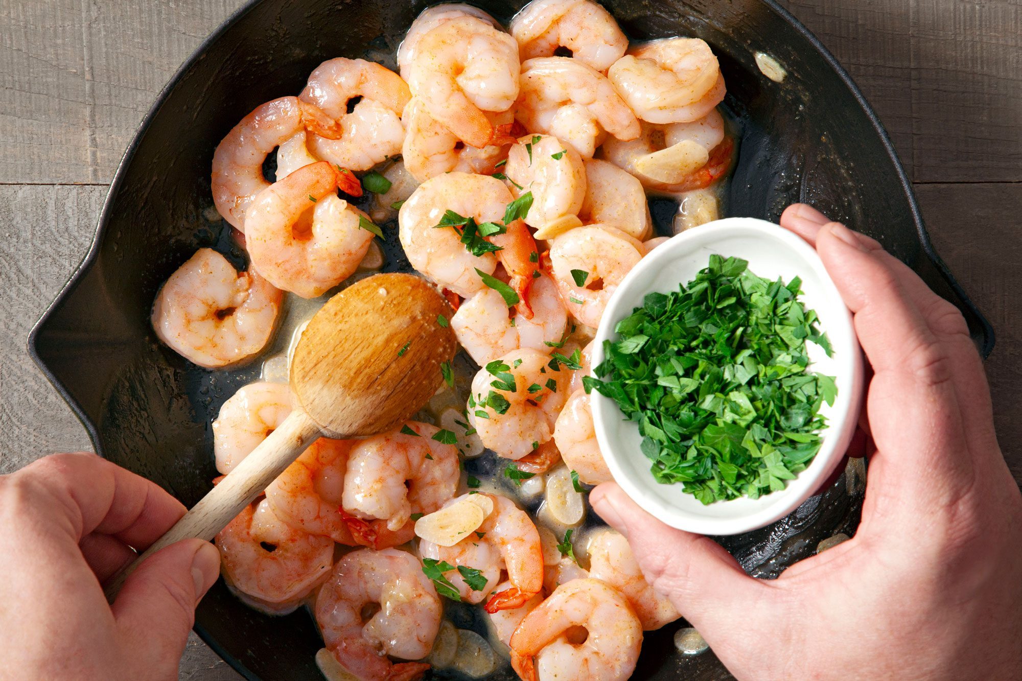 Overhead shot of cooked until shrimp turn pink; add parsley and stirred; wooden spoon; a small bowl of chopped parsley; wooden background;