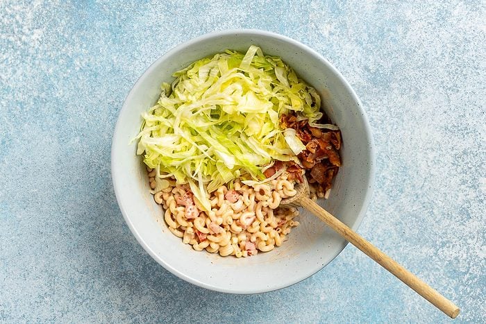 overhead shot of shredded lettuce and bacon added to the pasta salad mixture in a large serving bowl