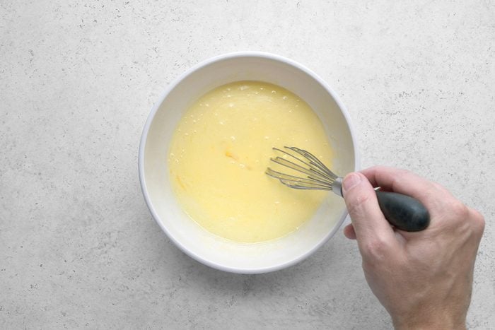 A person's hand whisking a yellow batter in a white bowl placed on a light gray countertop. The whisker has a black handle and the batter appears smooth. The scene suggests preparation for baking or cooking.