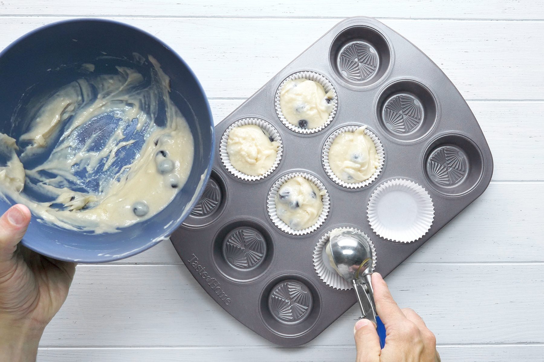 A person holding a blue mixing bowl with batter in one hand and a metal ice cream scoop in the other, filling a muffin tin lined with white paper cups on a white surface. 