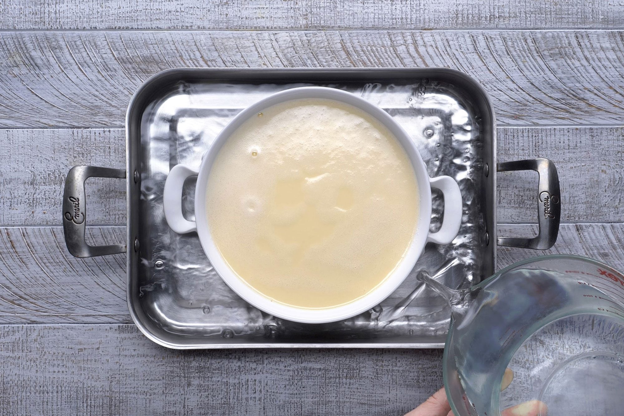 Overhead shot of place round baking dish in a larger baking pan; place pan on oven rack; add very hot water to pan to within 1/2 inch of top of round baking dish; bake until a knife inserted near the center comes out clean; wooden background;