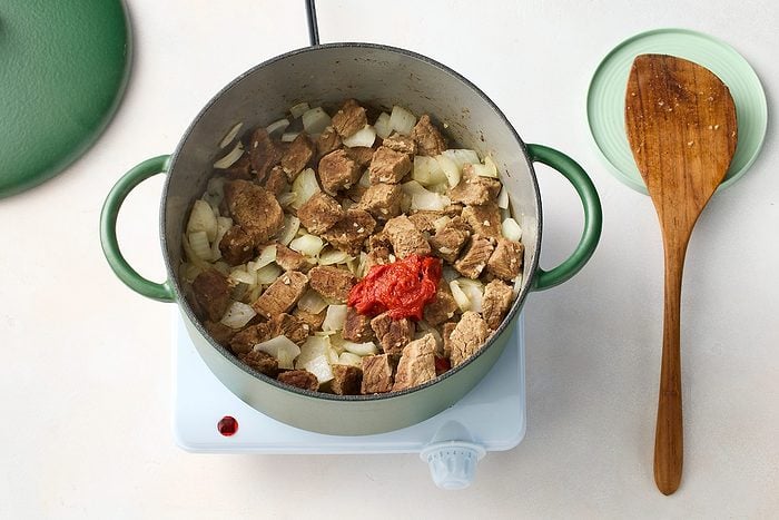 A green pot filled with chunks of beef, chopped onions, and a dollop of tomato paste is placed on a white electric stove.