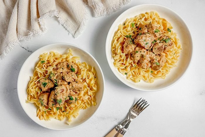 Taste of Home pork stroganoff in white ceramic bowls with a cloth napkin on a marble surface.
