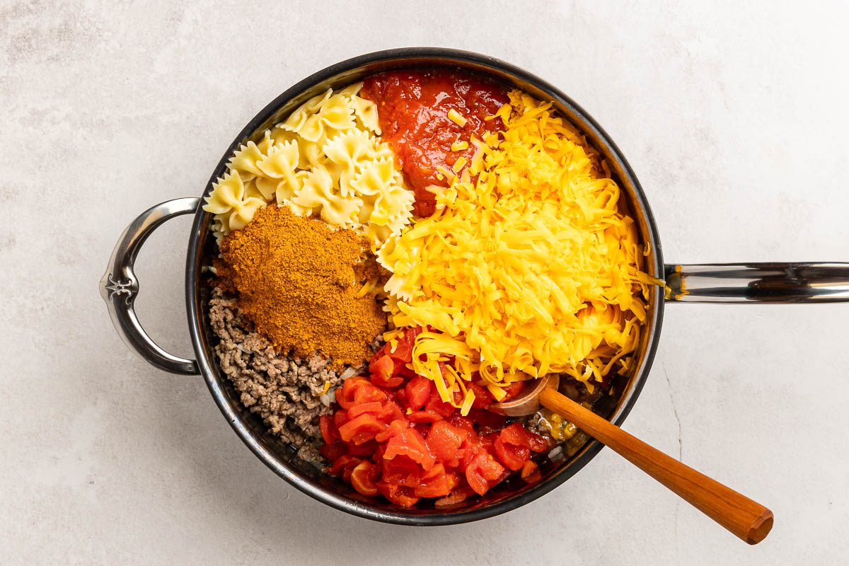 Overhead shot of cooked pasta, onion,crumbled beef,cheese, salsa, tomatoes and taco seasoning in a large skillet