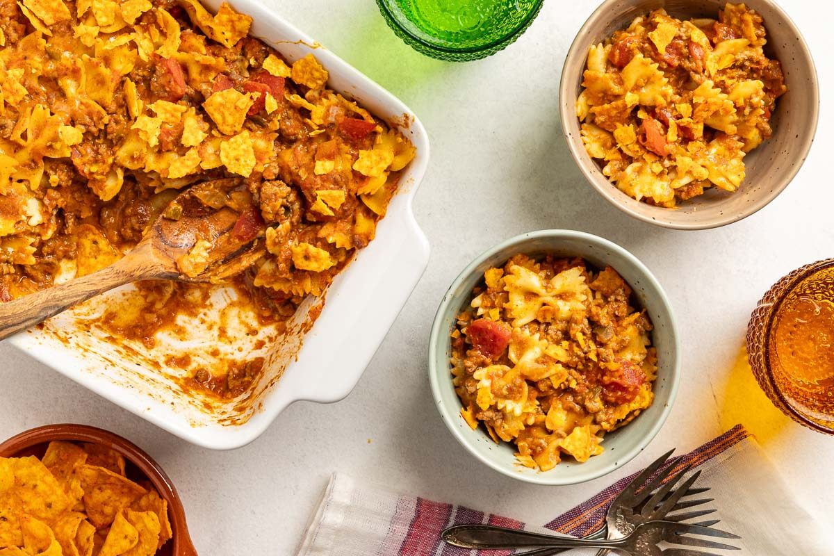 Overhead shot of Taco Casserole in a baking pan and served in bowls with forks, drinks, napkin and wooden spoon