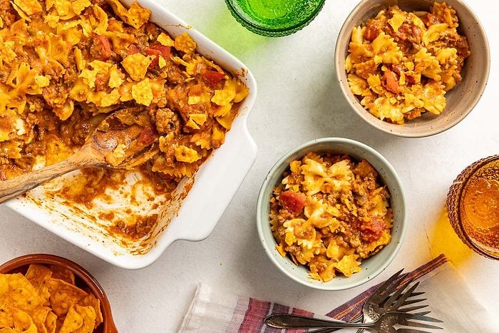 Overhead shot of Taco Casserole in a baking pan and served in bowls with forks, drinks, napkin and wooden spoon