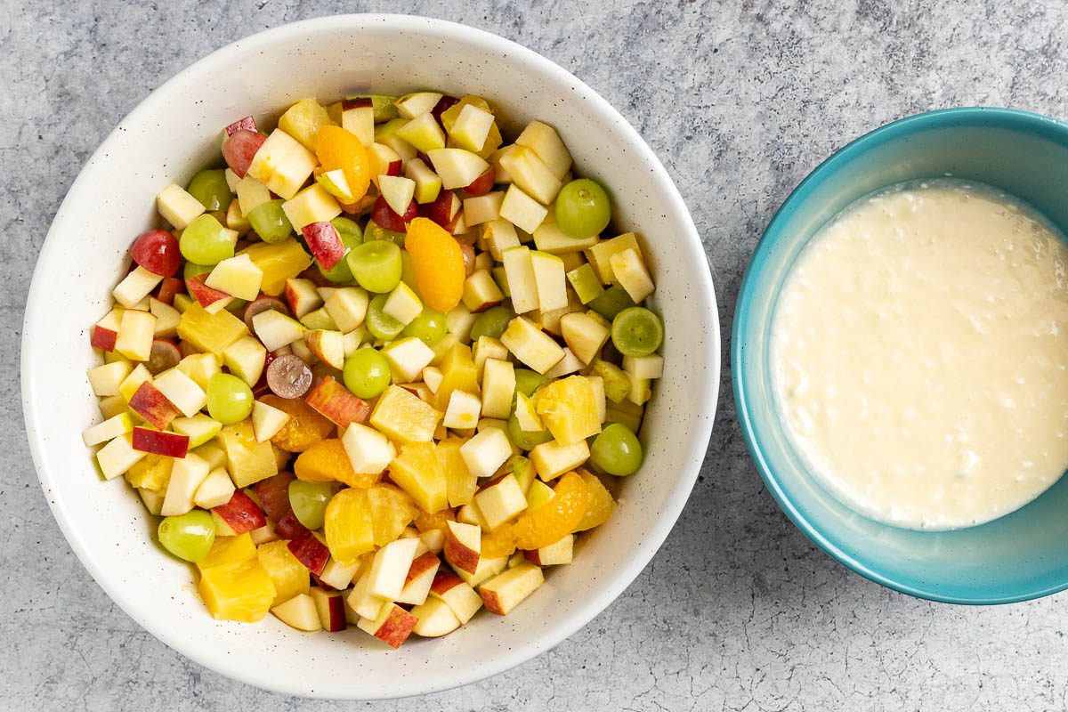 apples, grapes, pineapple chunks and mandarin oranges in a large bowl next to a smaller bowl of dressing