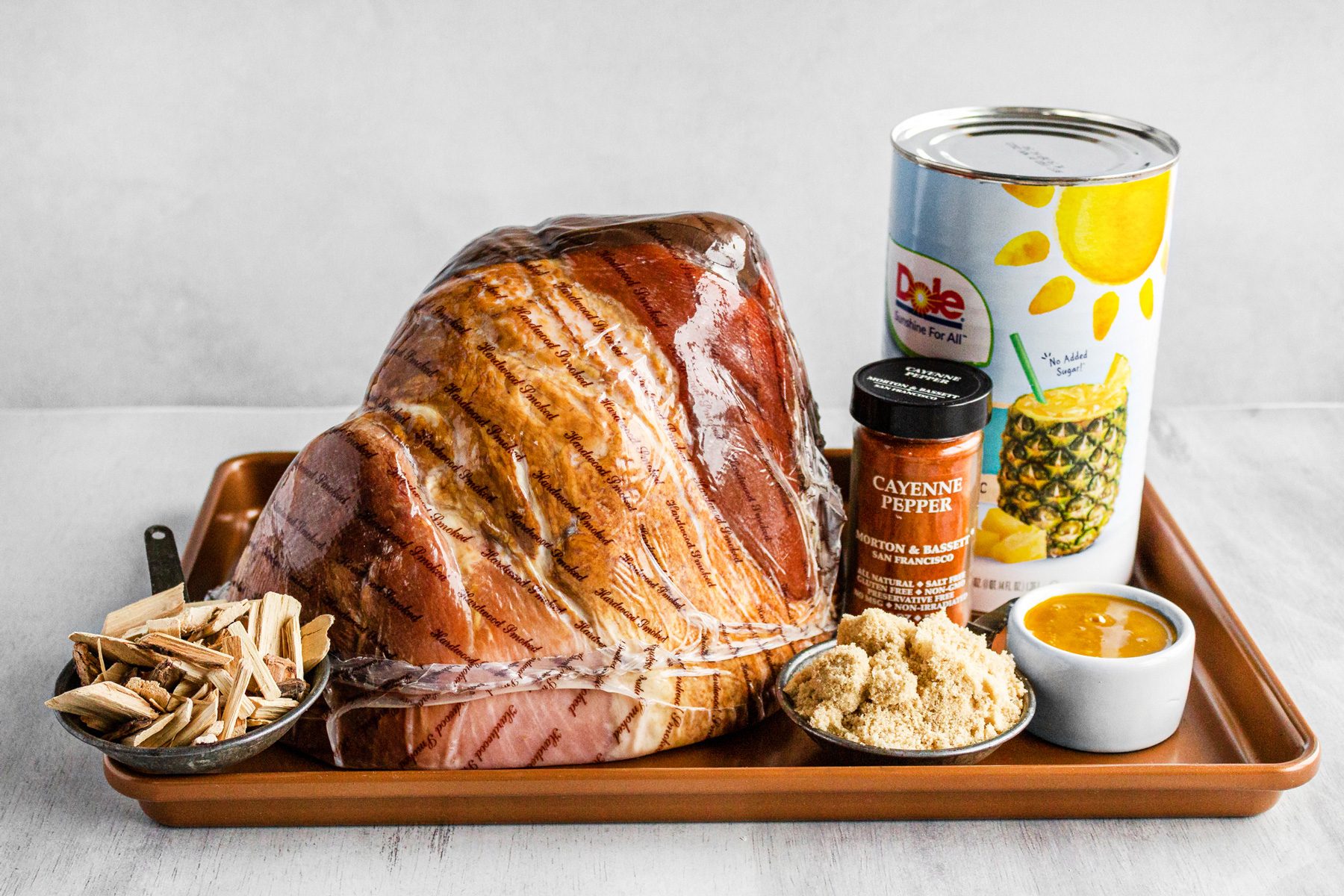 3/4th shot; white background; ham wrapped into transparent plastic; pineapple juice can; cayenne pepper jar bottle; in rectangular brown tray