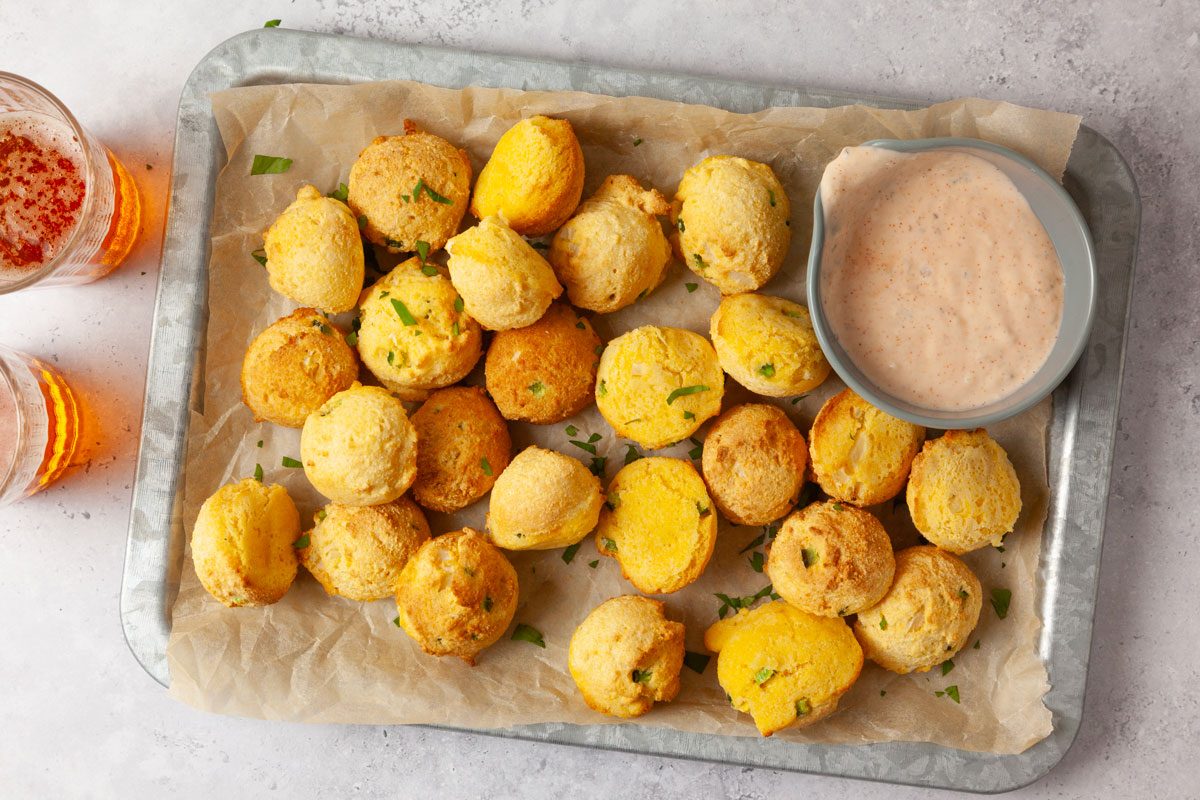 Full yield of Taste of Home Air-Fryer Hush Puppies on a metal tray lined with parchment paper, remoulade dipping sauce, beers