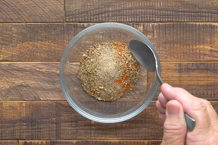 A person holding a spoon is mixing a variety of dried spices in a clear glass bowl. The bowl is placed on a wooden surface.