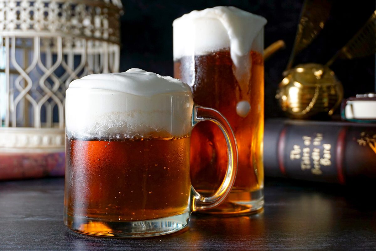 Two glass mugs filled with frothy butterbeer sit on a table. The drinks are amber with creamy foam on top. In the background, there is a book and decorative items,