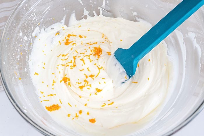 A glass bowl filled with a creamy white mixture is being stirred with a blue spatula. Orange zest is scattered on top of the mixture. The background is a light gray surface.