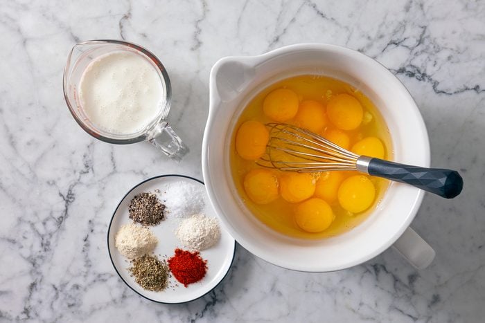 A white bowl contains several cracked eggs and a whisk. A small pitcher of milk sits nearby. A plate with various spices, including black pepper, salt, flour, oregano, and paprika, is also on the marble countertop.