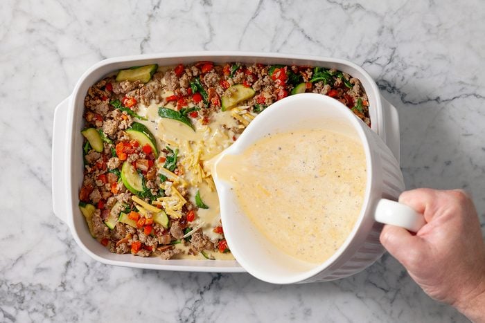 A person pours a creamy egg mixture over a casserole dish filled with ground meat, zucchini slices, chopped peppers, and spinach on a marble countertop.