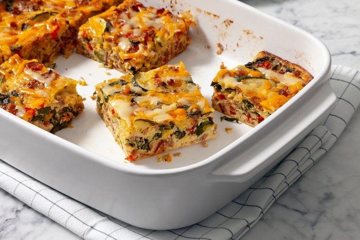 A white baking dish with four square servings of a colorful vegetable and cheese casserole. The dish rests on a checkered cloth on a marble surface. The casserole includes ingredients like zucchini, peppers, and melted cheese.
