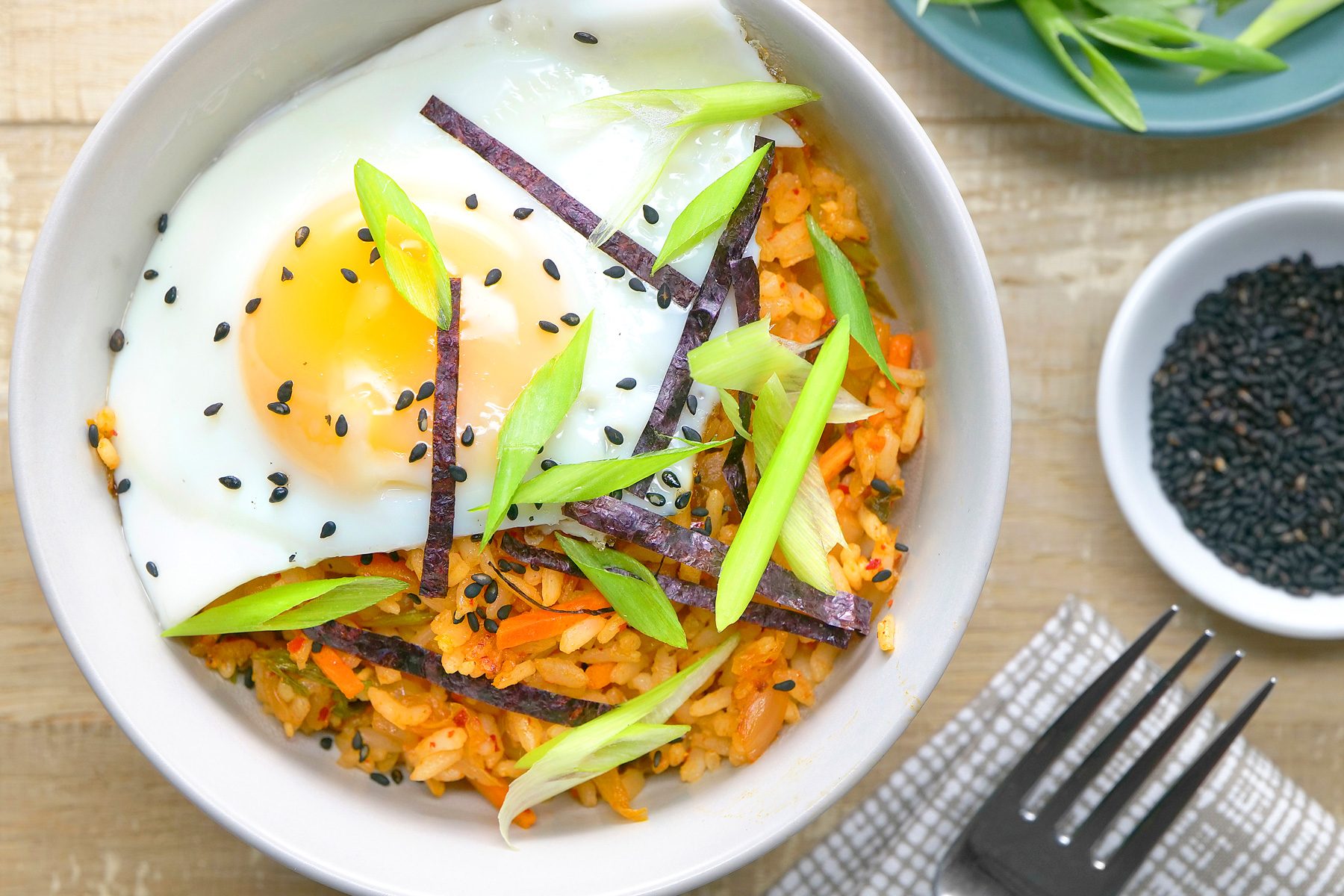 overhead shot of a bowl of Kimchi Fried Rice, The rice is tinged with a reddish hue from the kimchi and is accompanied by vibrant vegetables like carrots and green onions, A fried egg with a runny yolk sits atop the rice;