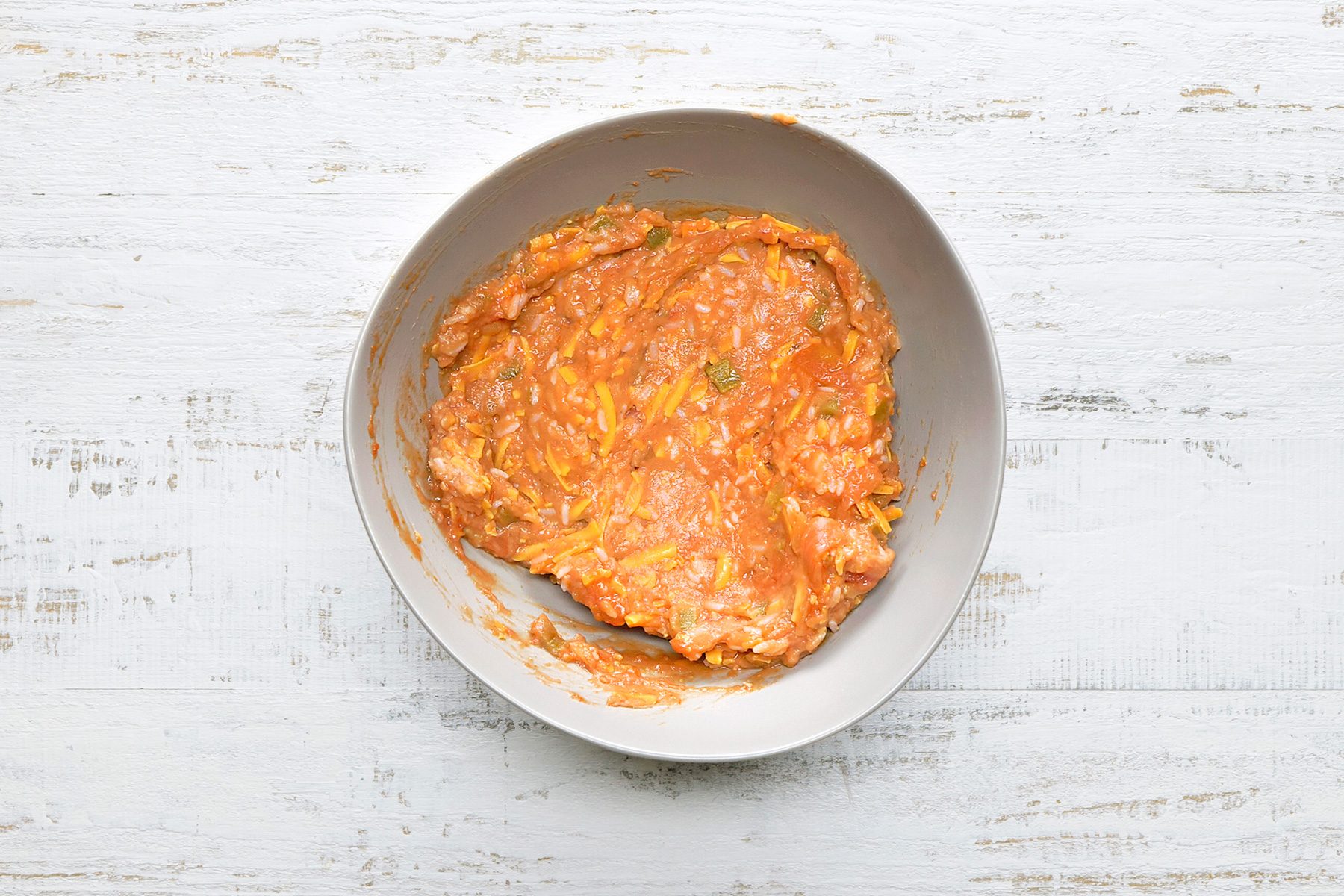 A gray bowl filled with a mixture of ground meat and vegetables, featuring a light red-orange hue with visible green herbs. The bowl is placed on a textured white wooden surface.