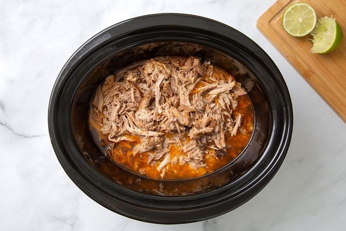 Shredded pork, pork juices, and mashed garlic in a slow cooker with juice added for step three of Chipotle Carnitas recipe for Taste of Home