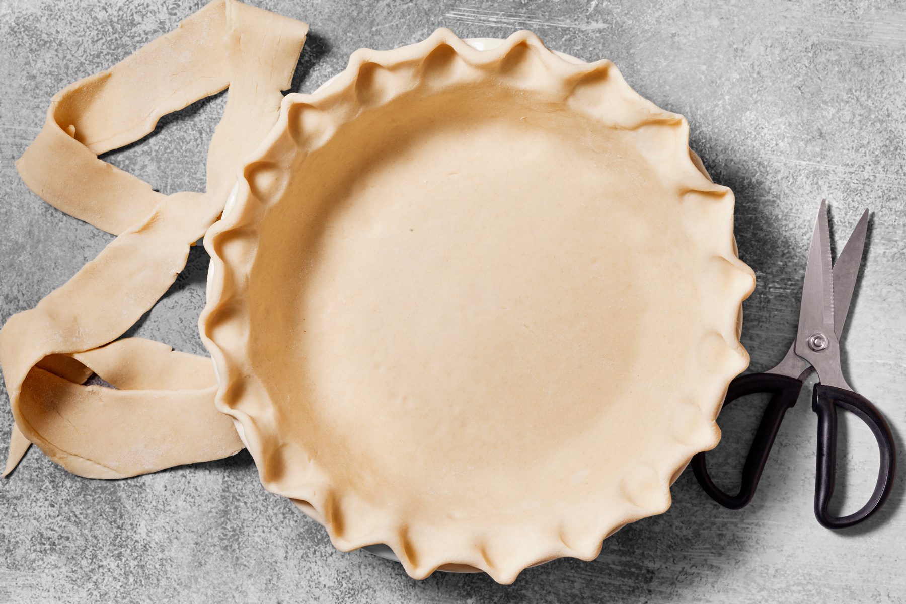 overhead shot of a pie crust that has been pressed into a pie pan, the crust has a fluted edge, the crust is light brown in color and there is some extra dough that was used to make the crust that is lying next to the pan, there is a pair of scissors next to the pie pan.